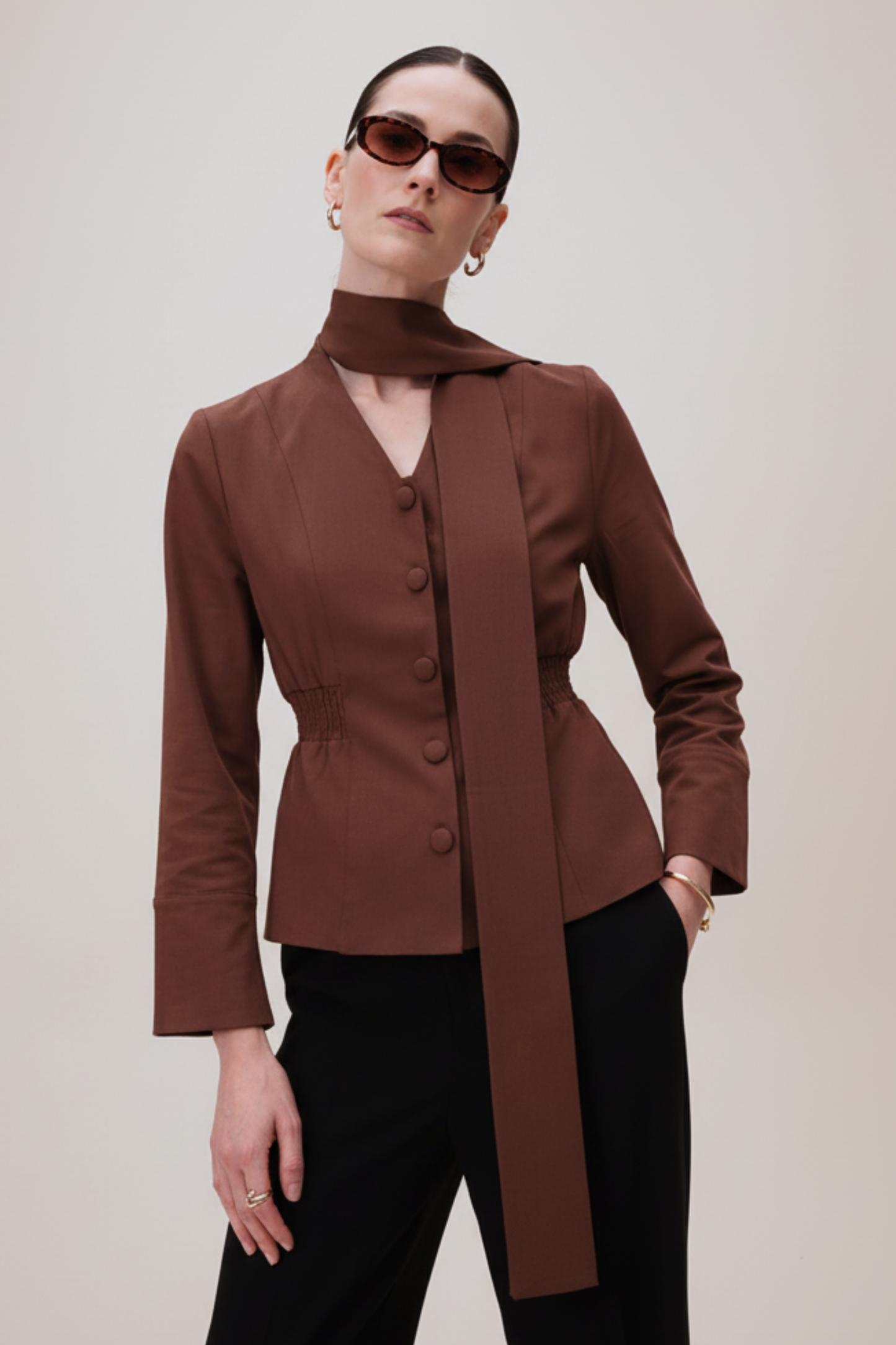Woman wearing a brown blazer with a long tie and sunglasses on a plain background
