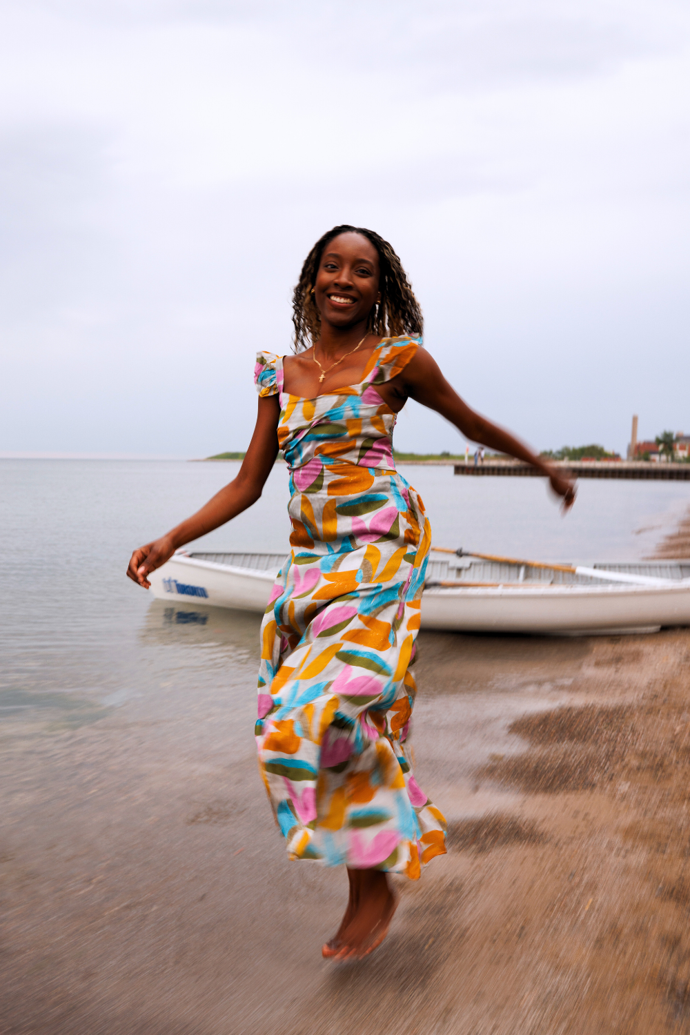 A woman twirling joyfully in a vibrant abstract-patterned dress near the shoreline with a boat and pier in the background.
