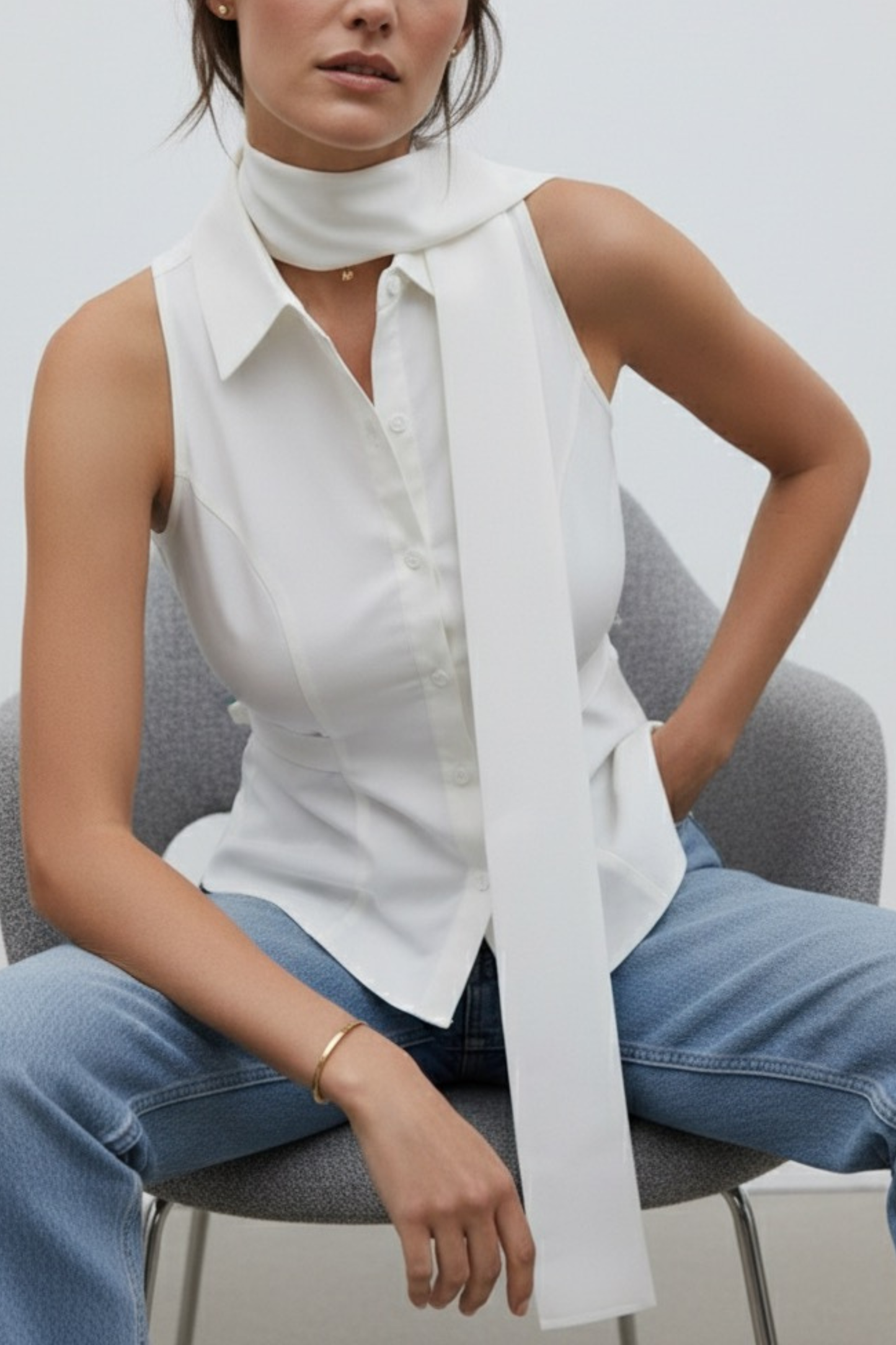 Woman wearing a white sleeveless blouse and blue jeans with a white scarf, sitting on a gray chair.