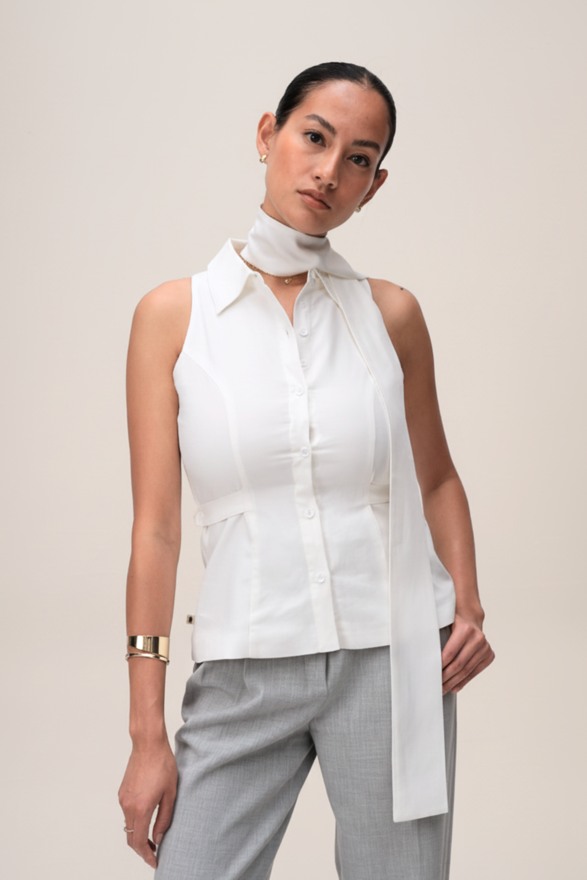 Woman wearing a white sleeveless top with a high collar and light gray pants on a beige background